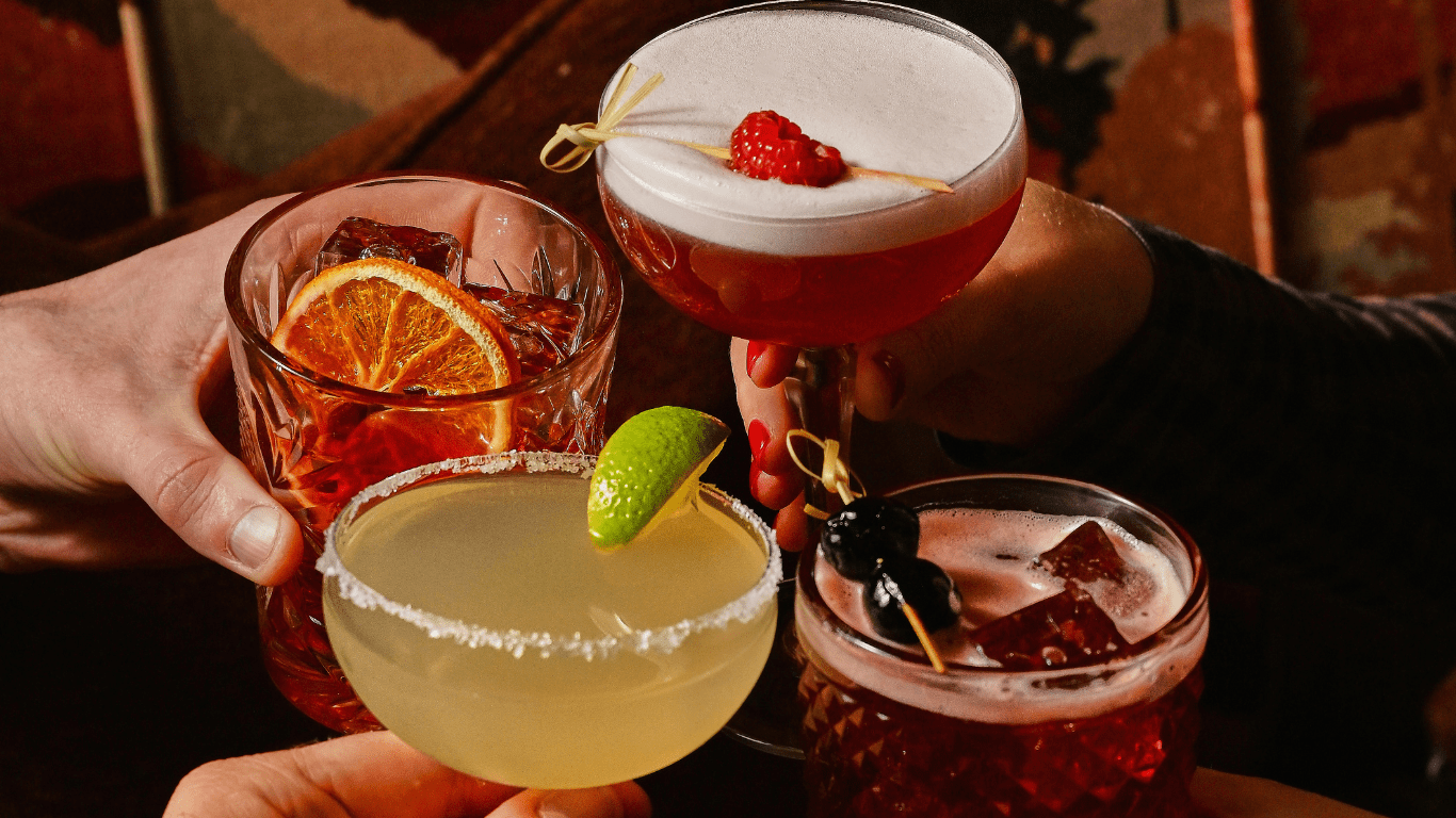 Four hands raise different cocktails: one with a dried orange slice, one with a lime wedge and salted rim, one with cherries and ice, and one with foam and raspberries, against a warm, dimly lit background.