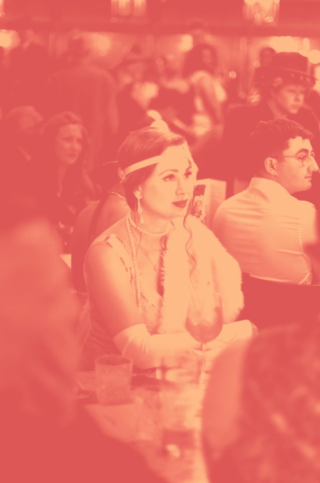 A woman in vintage 1920s attire, including pearl jewelry and a headband, sits at a table in a crowded, dimly lit event. The image has a pink tint and a soft, blurred effect.